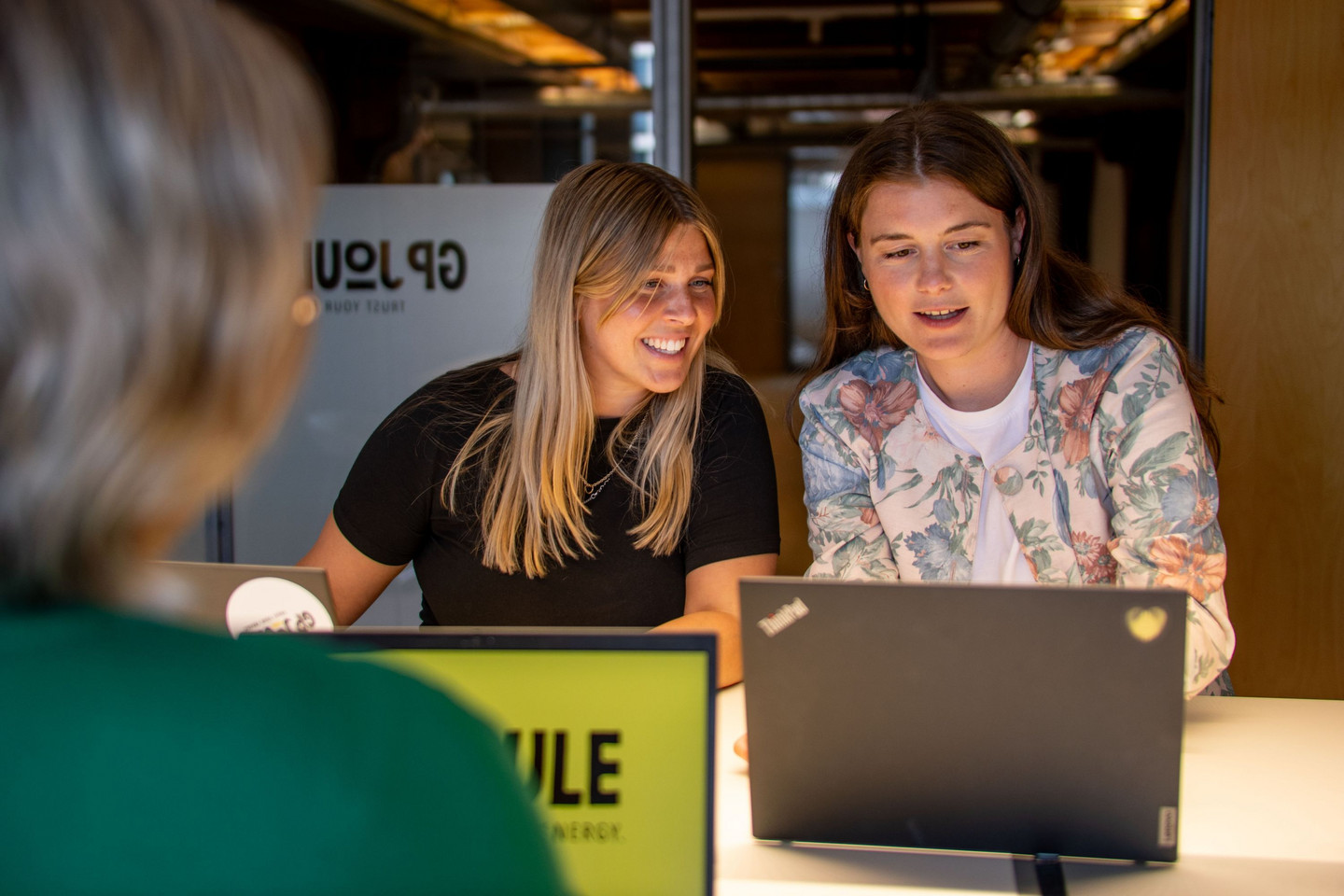 Zwei Frauen sitzen lächelnd mit Laptops am Tisch und arbeiten gemeinsam an etwas, eine dritte Person sitzt ihnen gegenüber.